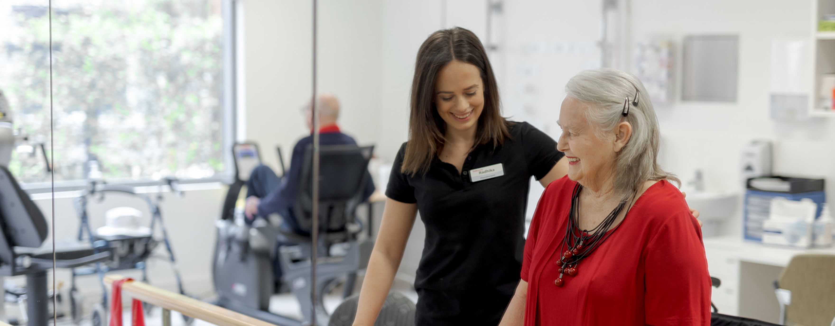 Eldercare physiotherapist Radhika Dick with Eldercare Allambi resident Bev Young at a set of parallel bars and smiling.