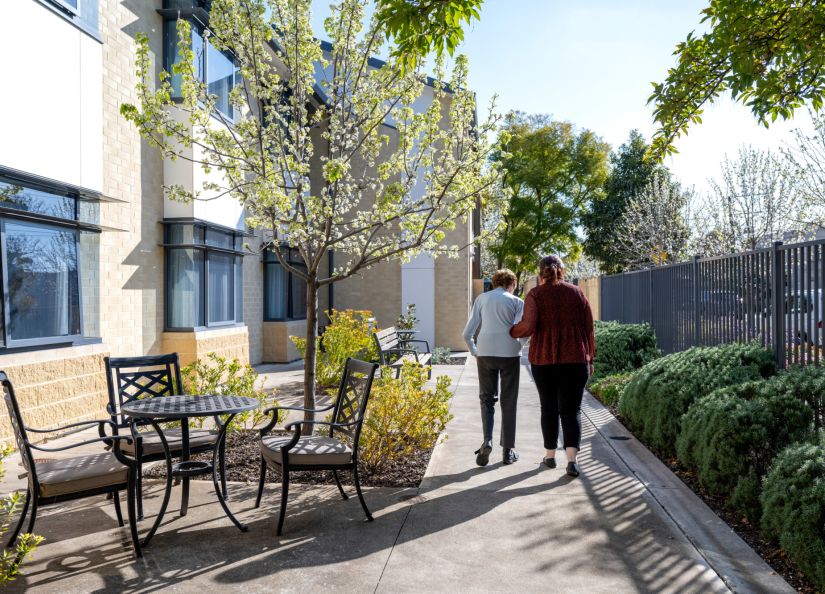 An Eldercare resident and volunteer walk through a sun-soaked courtyard at an aged care home.