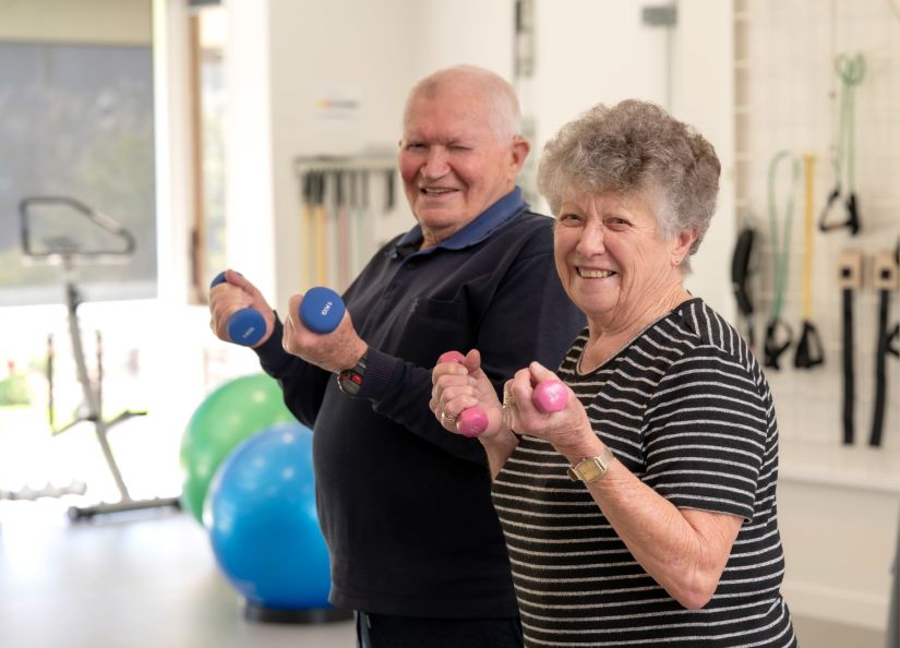 Two day therapy clients, a married couple, hold two dumbbells each and smile into the camera.